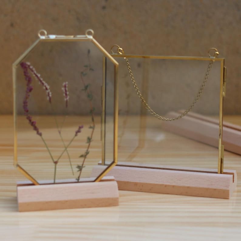 May include: Two gold-toned glass picture frames with wooden bases. The frame on the left is octagonal and contains dried flowers. The frame on the right is square and has a chain hanging from the top.