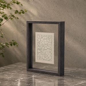 May include: A rectangular floating picture frame with a dark charcoal grey wooden border. Inside the clear glass panes, a small off-white rectangular paper features a continuous black line drawing of overlapping floral petal shapes.