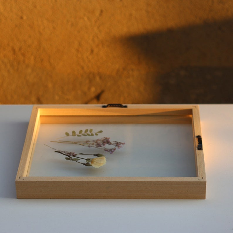 May include: A rectangular light-colored wooden floating frame containing pressed dried flowers. The frame features clear glass panes that sandwich a small green leaf, a sprig of pink baby's breath, and a single pale yellow flower blossom.