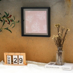 Puede incluir: Una impresión enmarcada en negro con un fondo rosa y un diseño floral blanco cuelga de una pared texturizada. Un calendario de madera muestra "DIC 29" debajo. También se ve un jarrón de vidrio transparente con flores secas y un libro.