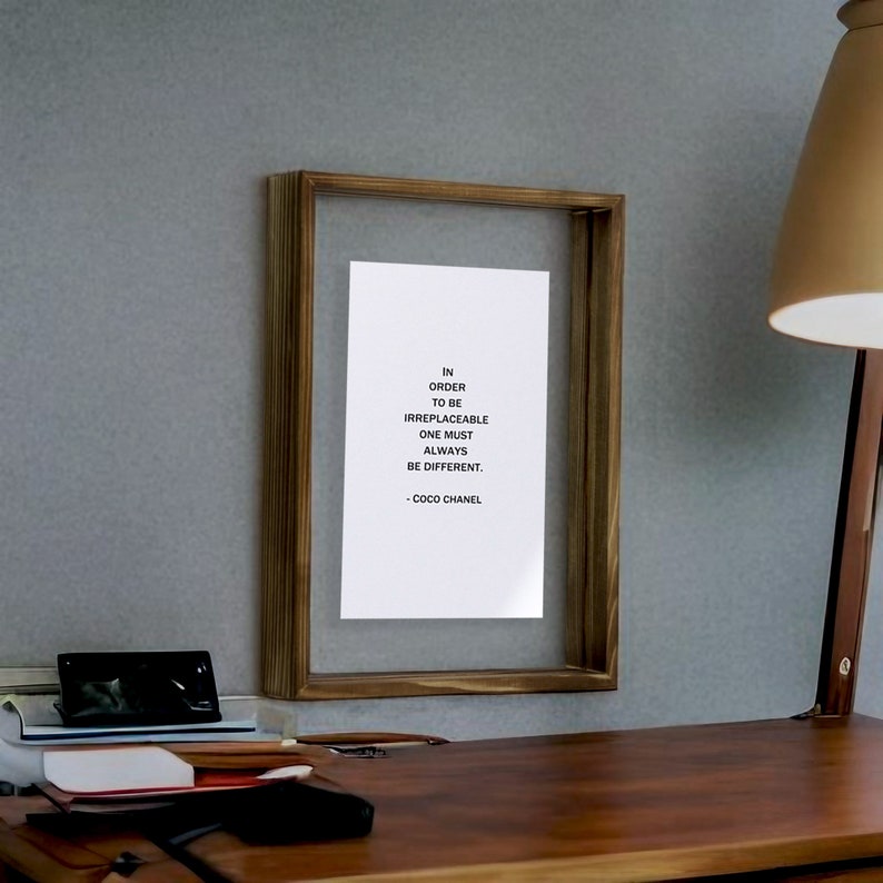 May include: A framed print with a quote by Coco Chanel: "In order to be irreplaceable one must always be different." The frame is brown wood and the print is on white paper.