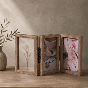 May include: A triptych photo frame made of light brown wood with a textured grain. The frame consists of three hinged vertical panels. The left panel holds a dried plant stem, the center panel displays a coffee cup and the text CELINE, and the right panel features a floral print with pink roses.