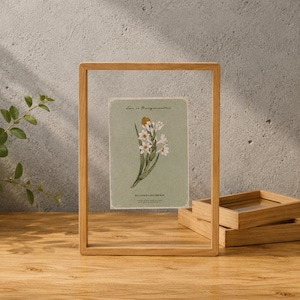 Può includere: Una cornice flottante rettangolare in legno chiaro che racchiude una carta verticale con sfondo verde salvia. La carta presenta un'illustrazione botanica di fiori bianchi con steli verdi e il testo Love is Unsymmetric stampato sopra.