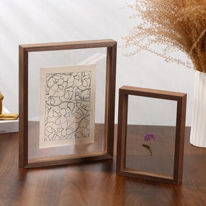 May include: Two rectangular floating picture frames made of dark brown wood. The larger frame contains a white paper insert with a black line drawing of floral shapes. The smaller frame holds a single pressed purple flower between the glass panes.