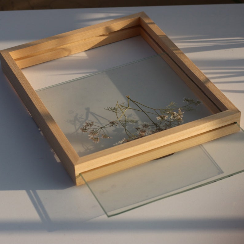 May include: A rectangular light-colored wooden floating frame featuring two clear glass panes. A delicate dried plant with small white flowers is pressed between the glass. One glass pane is partially slid out from the wooden frame structure.