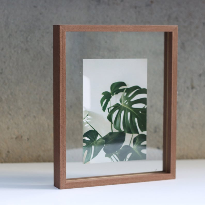 May include: A rectangular floating picture frame with a medium-toned brown wooden border. The frame features two clear glass panes holding a centered vertical print of a green monstera plant with large, split leaves against a white background.