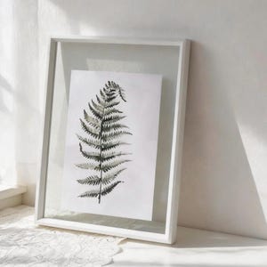 May include: A framed botanical print featuring a detailed fern illustration in shades of gray and black. The artwork is encased in a white, minimalist frame, creating a modern aesthetic. The print is displayed against a white background, enhancing the contrast.