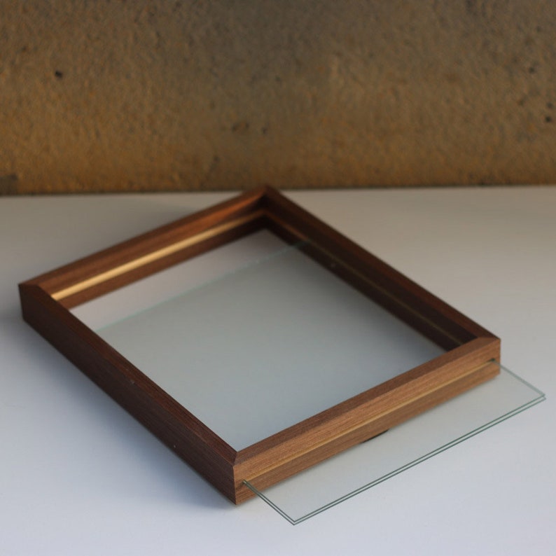 May include: A rectangular wooden picture frame with a medium brown finish and a visible wood grain texture. The frame features a clear glass pane that is partially slid out from the side, revealing the internal channel where the glass rests.