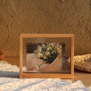 May include: A rectangular light-coloured wooden floating picture frame with a clear glass centre. Inside the frame is a photograph of a woven basket filled with white flowers and green stems, resting against a soft, light-coloured fabric background.