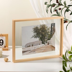May include: A rectangular floating picture frame with a light-colored wooden border containing a print of a Mediterranean coastal path with white walls, a large tree, and a distant sea view, displayed in a clear glass panel design.