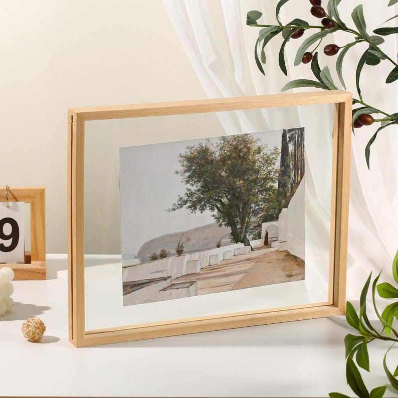 May include: A rectangular floating picture frame with a light-colored wooden border containing a print of a Mediterranean coastal path with white walls, a large tree, and a distant sea view, displayed in a clear glass panel design.