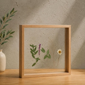 May include: A square wooden floating frame featuring two clear glass panes. Inside the frame, several pressed botanical elements are displayed, including green leaves, a purple flower spike, thin stems, and a single white daisy-like flower.