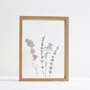May include: A wooden frame with a clear glass front displays pressed purple and white flowers. The flowers are arranged in a simple, natural design.