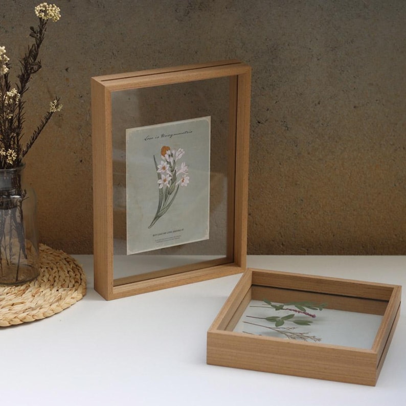 May include: Two wooden picture frames with glass fronts. The larger frame is vertical and holds a botanical illustration of white flowers. The smaller frame is square and holds a pressed flower arrangement.
