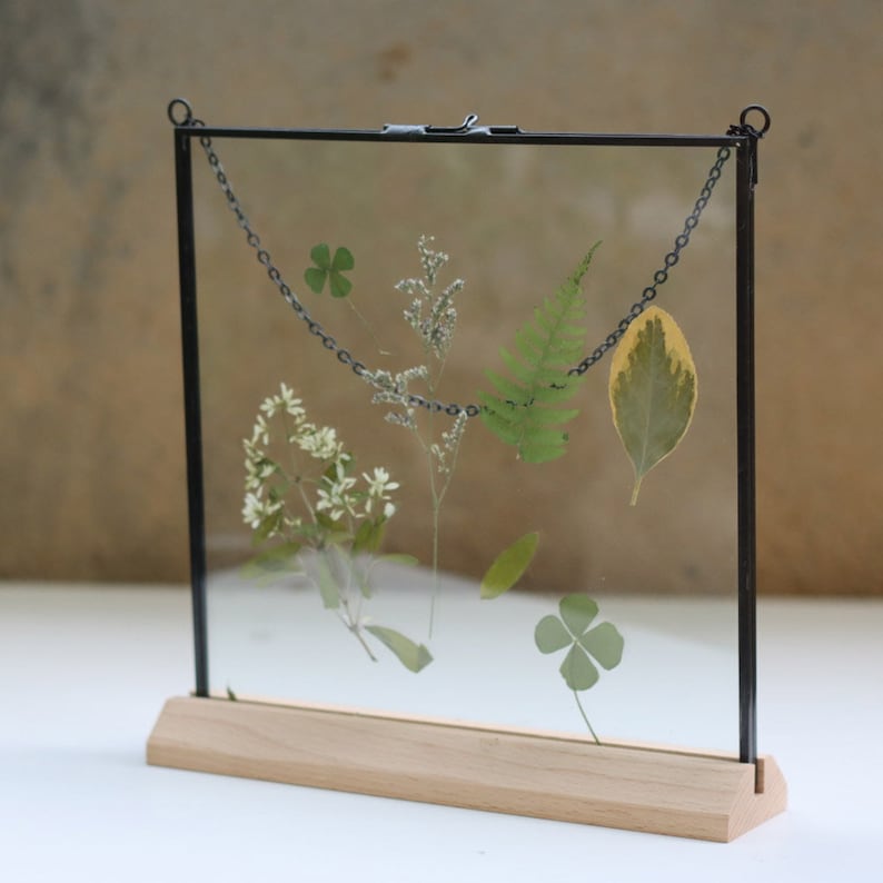 May include: A black metal frame with a wood base holds pressed flowers and leaves. The frame has a chain hanging from the top.