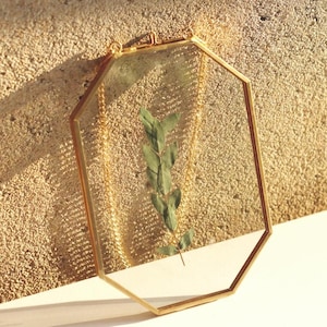 May include: A gold octagonal picture frame with a glass front. The frame is hanging from a chain and has a sprig of dried greenery inside.