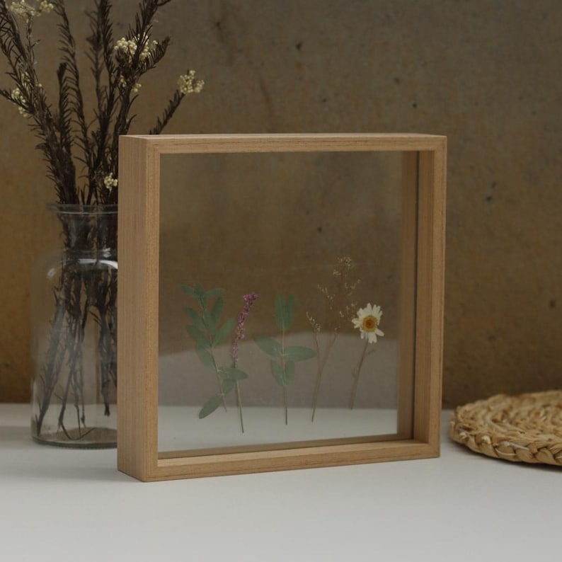 May include: A square wooden frame with a clear glass front, showcasing pressed flowers and greenery. The frame is light brown and has a simple, minimalist design.