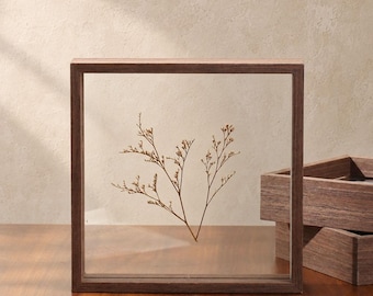 May include: A square wooden frame with a dark brown finish containing two clear glass panes. A single dried, light brown sprig of statice is pressed between the glass. A second, empty wooden frame is stacked partially behind the first frame.