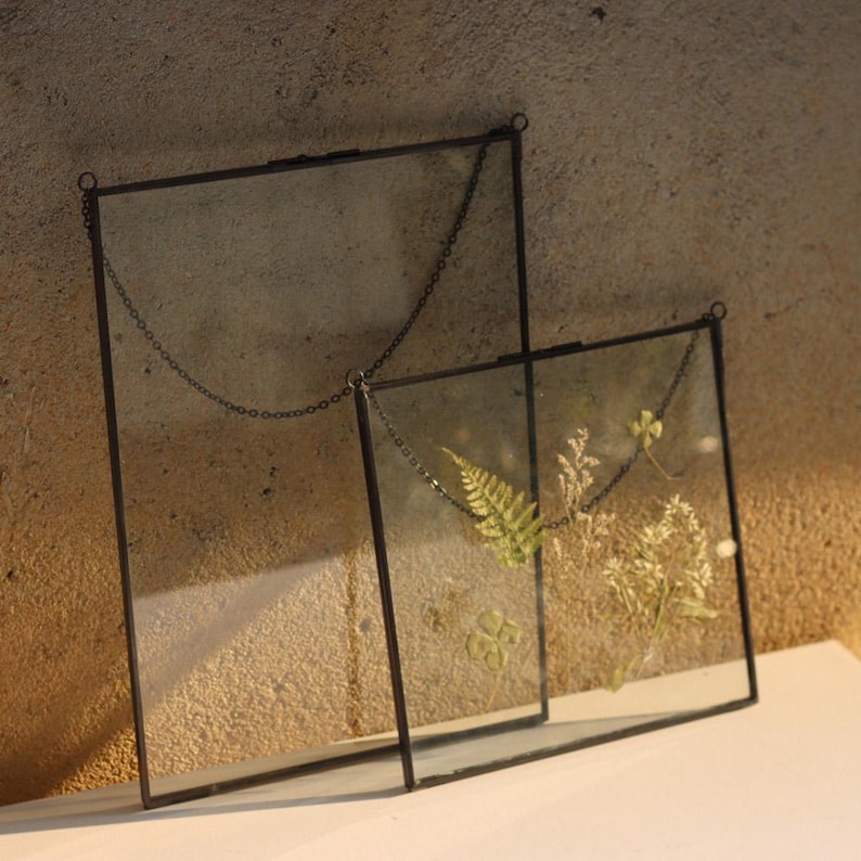 May include: Two black metal picture frames with glass panels, hanging from chains. The smaller frame contains pressed flowers and ferns.