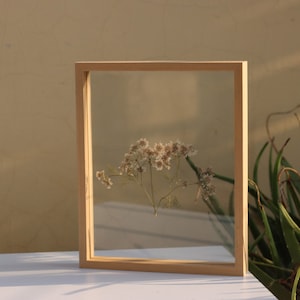 May include: A rectangular light-colored wooden floating frame containing a single pressed sprig of small white flowers. The frame features two clear glass panes held within a simple, thin, natural wood border, showcasing the delicate botanical specimen.
