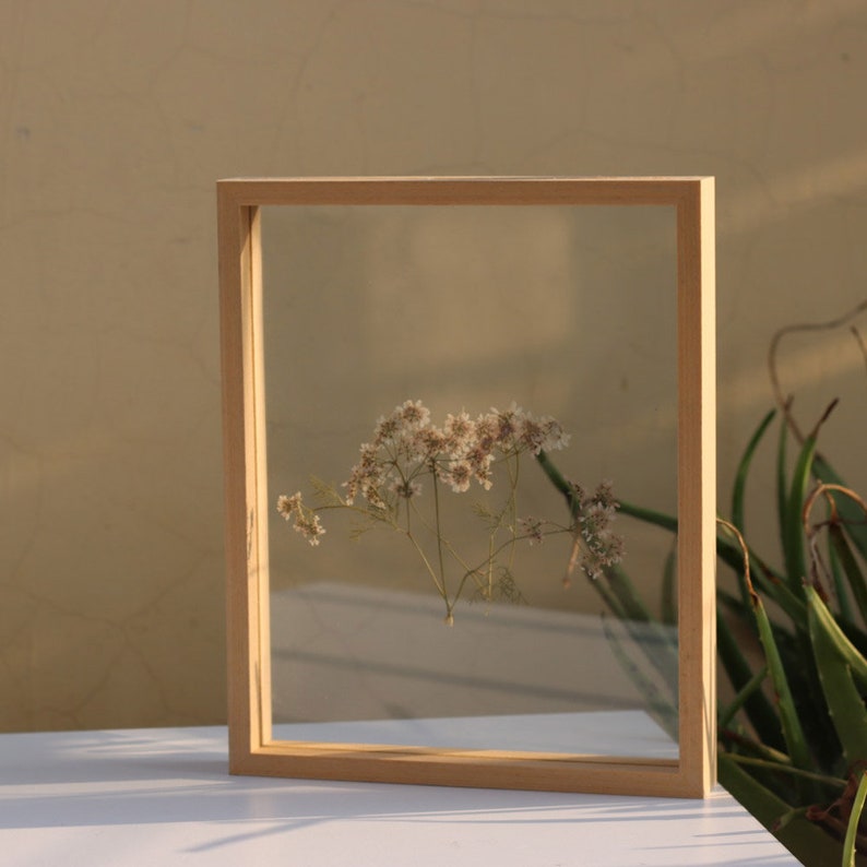 May include: A rectangular light-colored wooden floating frame containing a single pressed sprig of small white flowers. The frame features two clear glass panes held within a simple, thin, natural wood border, showcasing the delicate botanical specimen.