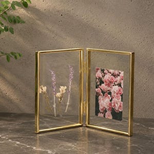May include: A double-hinged gold metal picture frame with clear glass panels. The left side holds pressed dried purple and white flowers, while the right side displays a rectangular photograph of pink rose blossoms in a dense cluster.