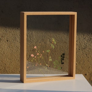 Op de afbeelding: Een vierkant houten frame met een heldere glazen inzet, met geperste bloemen en bladeren. Het frame is lichtbruin en heeft een natuurlijke houten afwerking.