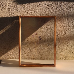 May include: A wooden frame with a clear glass insert, showcasing a dried flower arrangement. The frame is a light brown colour and has a rustic appearance. The flower is a light purple colour and is positioned in the centre of the frame.