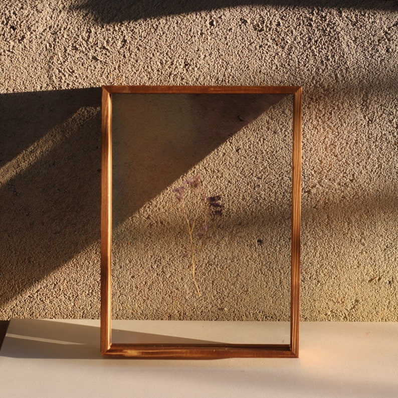 May include: A wooden frame with a clear glass insert, showcasing a dried flower arrangement. The frame is a light brown colour and has a rustic appearance. The flower is a light purple colour and is positioned in the centre of the frame.