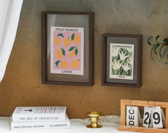 May include: Two rectangular dark wood frames containing prints. The left print features yellow lemons on a pink background with the text FRUIT MARKET LEMON. The right print shows white flowers on a green background with the text FLOWER MARKET TOKYO.