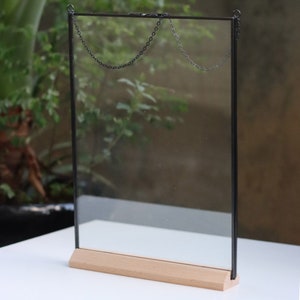 May include: A black metal frame with a clear glass panel and a wooden base. The frame has two chains attached to the top for hanging.