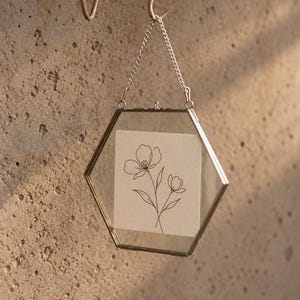 May include: A hexagonal glass frame with a thin metallic border, suspended by a silver-toned chain. Inside the transparent panes, a square off-white card features a black line art illustration of two delicate flowers with stems and leaves.