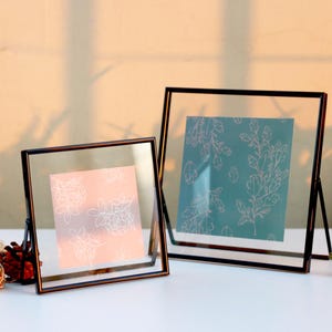 May include: Two square glass photo frames with black metal borders. One frame holds a pink print with white floral designs, the other a teal print with white botanical patterns. Both frames are displayed on a white surface.