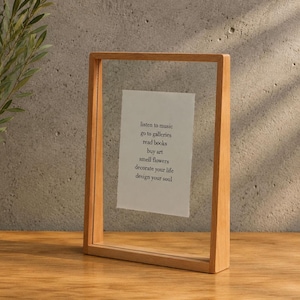 Op de afbeelding: Een rechthoekige, zwevende fotolijst van licht hout met twee heldere glasplaten die een wit papieren inzetstuk vasthouden. De tekst op het papier luidt: listen to music, go to galleries, read books, buy art, smell flowers, decorate your life, design your soul.