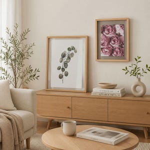 May include: Two framed wall art pieces featuring a watercolor eucalyptus branch and a cluster of pink peonies, displayed on a light wood media console alongside a beige ceramic vase, a wooden bowl, and two stacked books titled The Kinfolk Home.