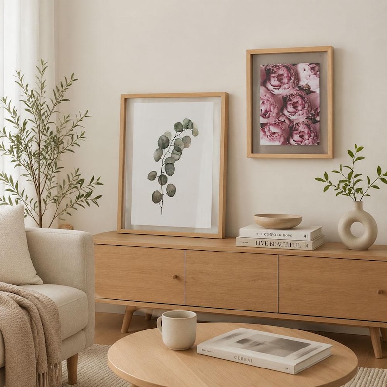 May include: Two framed wall art pieces featuring a watercolor eucalyptus branch and a cluster of pink peonies, displayed on a light wood media console alongside a beige ceramic vase, a wooden bowl, and two stacked books titled The Kinfolk Home.
