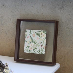May include: A square floating frame with a dark brown wooden border containing a piece of paper with a botanical pattern. The print features green fern leaves, white flowers, and small pink blossoms set against a light sage green background.