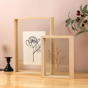 Puede incluir: Dos marcos de madera con cristal. Uno contiene un dibujo lineal negro de una rosa sobre papel crema. El otro contiene ramitas secas de color marrón claro. Un candelabro negro está en el fondo.