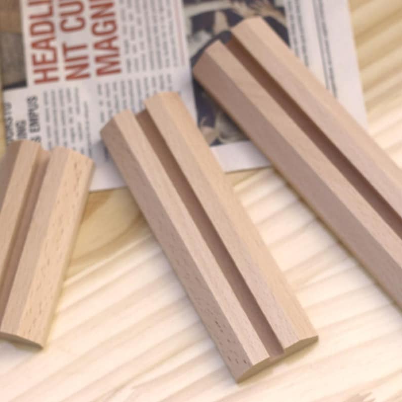 May include: Three wooden display stands with a groove in the center. The stands are made of light brown wood and are arranged on a light wood surface.