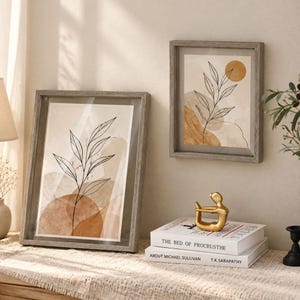 May include: Two framed wall art prints featuring black line drawings of leafy branches over abstract tan and beige watercolor shapes. The frames are made of gray wood. A gold abstract figurine sits on two stacked books titled The Bed of Procrusthe.