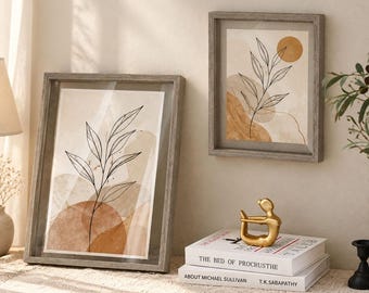 Könnte beinhalten: Zwei gerahmte Wandbilder mit schwarzen Strichzeichnungen von Blattzweigen auf abstrakten beigen und ockerfarbenen Aquarellformen. Die Rahmen bestehen aus grauem Holz. Eine goldene abstrakte Figur steht auf zwei Büchern mit dem Titel The Bed of Procrusthe.