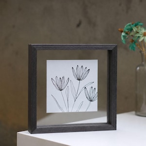 May include: A square floating frame with a dark gray wood finish containing a white insert with a black line drawing of three stylized flowers with pointed petals and long stems, creating a simple botanical illustration within the glass panes.