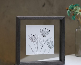 Puede incluir: Un marco flotante cuadrado con acabado en madera gris oscuro que contiene un inserto blanco con un dibujo de líneas negras de tres flores estilizadas con pétalos puntiagudos y tallos largos, creando una ilustración botánica simple.