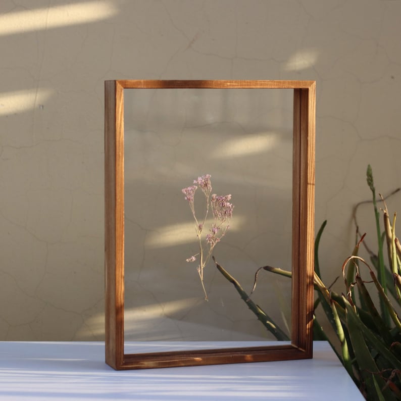 May include: A wooden frame with a clear glass insert, showcasing a dried flower arrangement with delicate pink blossoms. The frame is standing on a white surface.