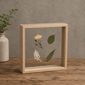 May include: A square light-colored wooden floating frame containing four pressed botanical specimens: a pale yellow flower, a serrated green leaf, a white dusty miller leaf, and a green fern frond, all displayed between two clear glass panes.