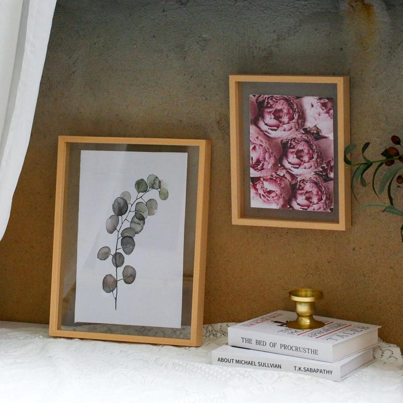 May include: Two light-colored wooden picture frames. One larger frame displays a watercolor-style illustration of a green eucalyptus branch. The smaller frame contains a print of pink peony flowers. A gold candle holder sits on two stacked books.