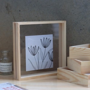 Puede incluir: Un marco de fotos de madera con un inserto de acrílico transparente, que muestra un dibujo de línea en blanco y negro de flores. El marco está hecho de madera natural y tiene un diseño simple y minimalista.