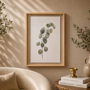 May include: A framed wall art piece featuring a watercolor illustration of a single eucalyptus branch with round, sage green and grey leaves on a white background, enclosed in a light-toned wooden frame with a clear glass front panel.