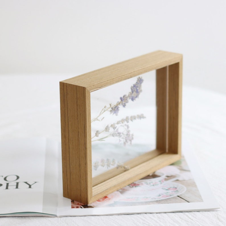May include: A wooden frame with a clear acrylic insert, showcasing dried purple flowers. The frame is sitting on a magazine with a floral pattern.