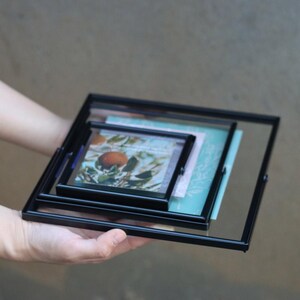 以下が含まれることがあります： 黒い金属とガラス製の正方形のフォトフレームが3つ重ねられています。それぞれのサイズは小さくなっています。フレームには画像と装飾用の紙が入っています。背景はニュートラルグレーです。